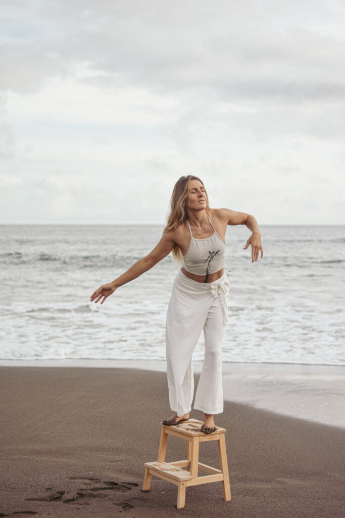 Yoga-Lehrerin in weißer Kleidung sitzt am Strand auf einem Hocker, macht feminine Bewegungen mit geschlossenen Augen – Embodiment, Weiblichkeit und fließende Energie.