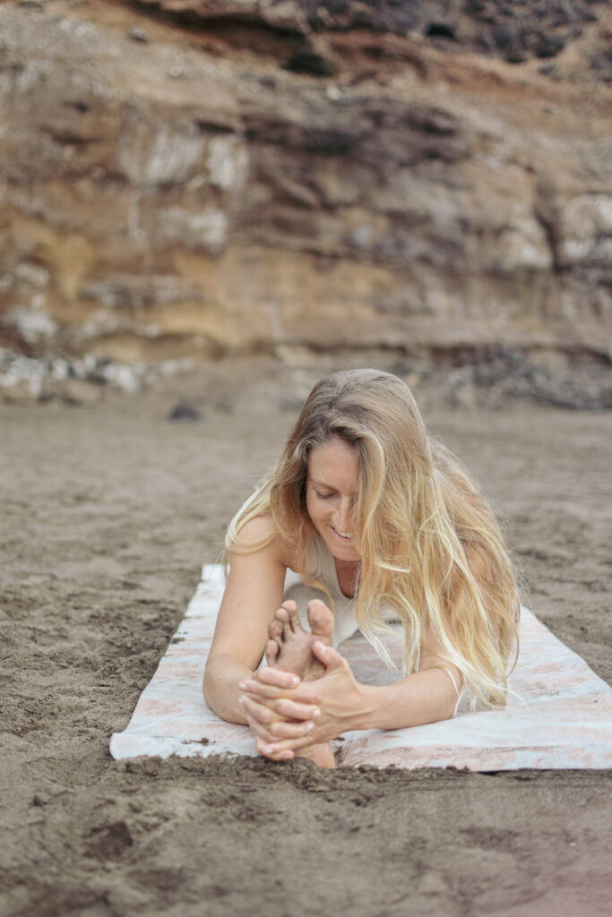 Yoga-Lehrerin auf Yogamatte im Sand vor Felsen, in einbeiniger Vorwärtsbeuge, lachend nach unten blickend – Yoga für den weiblichen Zyklus, Leichtigkeit und Körperbewusstsein.