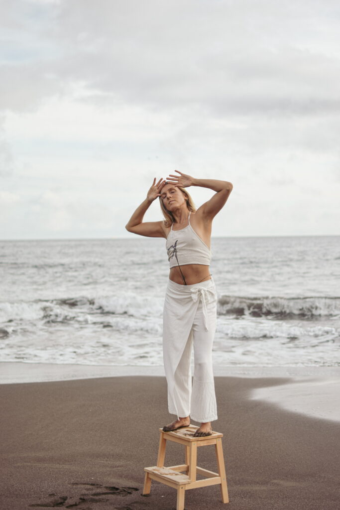 Yoga-Lehrerin in weißer Kleidung steht auf einem Hocker am Strand, Blume in der Hose, Hände an den Schläfen, Augen geschlossen, Gesicht nach oben gerichtet – Weiblichkeit, Naturverbundenheit und Selbstreflexion.
