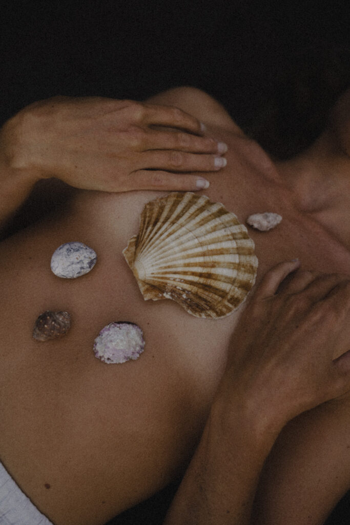 Frau mit Muscheln bedeckt, die Hände sanft auf der Brust, Oberkörper sichtbar. Das Bild strahlt Weiblichkeit, Selbstliebe und Verbundenheit mit der Natur aus.