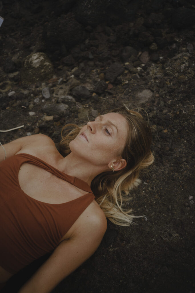 Frau in ockerrotem Yoga-Outfit liegt auf Steinen, der Blick bedacht in den Himmel gerichtet. Ausdruck von Erdung, innerer Reflexion und Verbindung zur Natur.