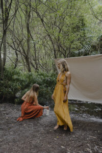 Zwei Frauen stehen barfuß im Dschungel, in langen fließenden Kleidern, und verkörpern weibliche Energie, zyklisches Leben und die Rückkehr zur Natur. Das Bild symbolisiert Verbindung, Embodiment und die Kraft des weiblichen Zyklus im Einklang mit der Natur.