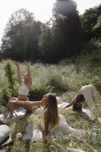 Frauen sitzen barfuß im Kreis auf einer Wiese, umgeben von Gras und Natur, halten sich an den Händen oder haben ihre Hände auf ihren Herzen, während sie miteinander verbunden sind. Das Bild strahlt Gemeinschaft, Weiblichkeit und die heilsame Kraft von Women’s Circles in der Natur aus.