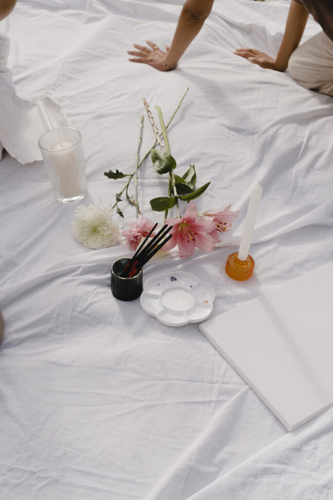 Picknickdecke ausgebreitet auf einer Wiese, darauf verteilt liegen Frauenutensilien wie Journals, eine Teekanne, Blumen, ein Ritualkerzenhalter und Kristalle, die eine warme, einladende Atmosphäre für zyklisches Leben, Selbstfürsorge und Frauenrituale im Alltag schaffen.